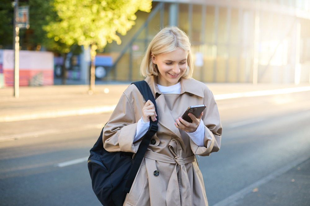 smiling-blonde-woman-trench-coat-standing-street-using-mobile-phone-app-holding-backpack.jpg